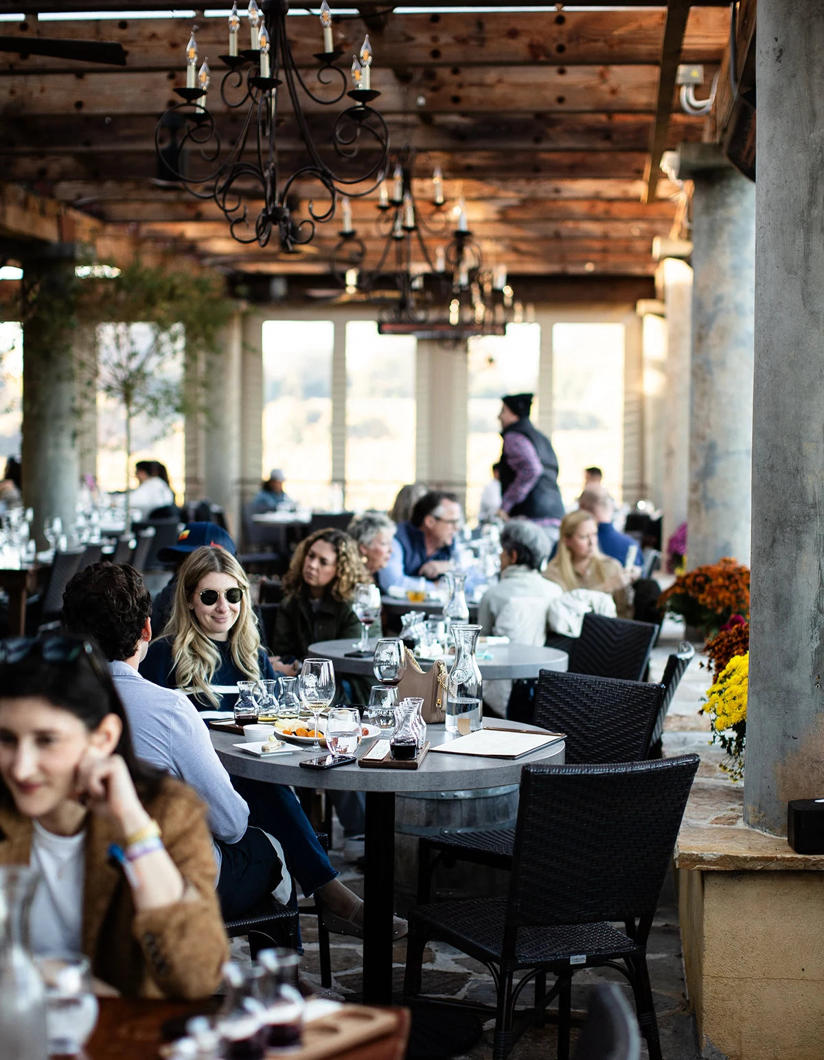 People dining in a rustic outdoor restaurant setting with wooden beams and large windows.