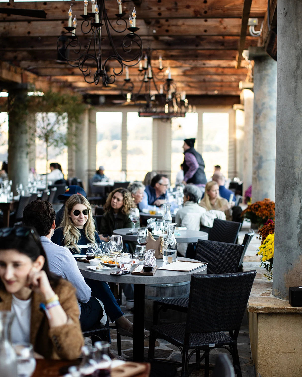 People dining in a rustic outdoor restaurant setting with wooden beams and large windows.